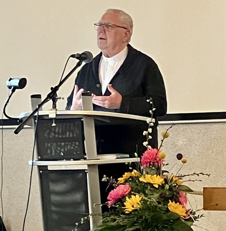 Diakon Helmut Zimmermann steht vorne im Saal und hält eine Predigt während der Gedenkfeier. Er spricht zu den Anwesenden in ruhiger, würdevoller Atmosphäre; im Hintergrund ist der Veranstaltungsraum mit Sitzreihen erkennbar. Die Szene vermittelt Besinnlichkeit, Trost und Hoffnung im Rahmen der Abschiedsfeier.
